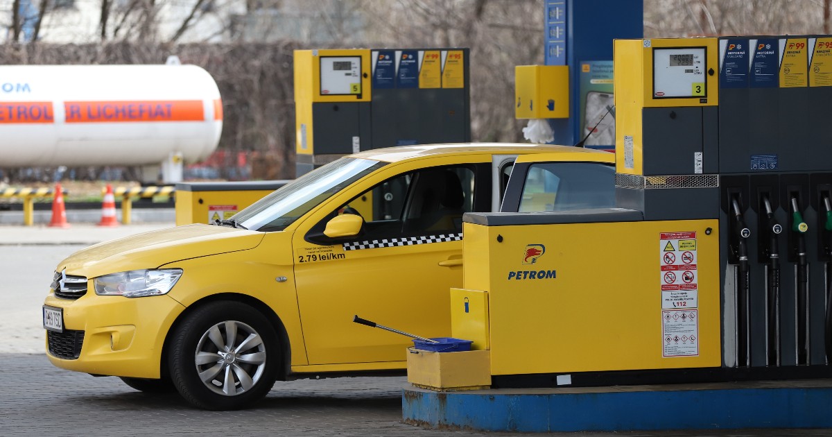 Imaginea prezintă un autoturism taxi galben staționat lângă o pompă de alimentare într-o benzinărie Petrom.