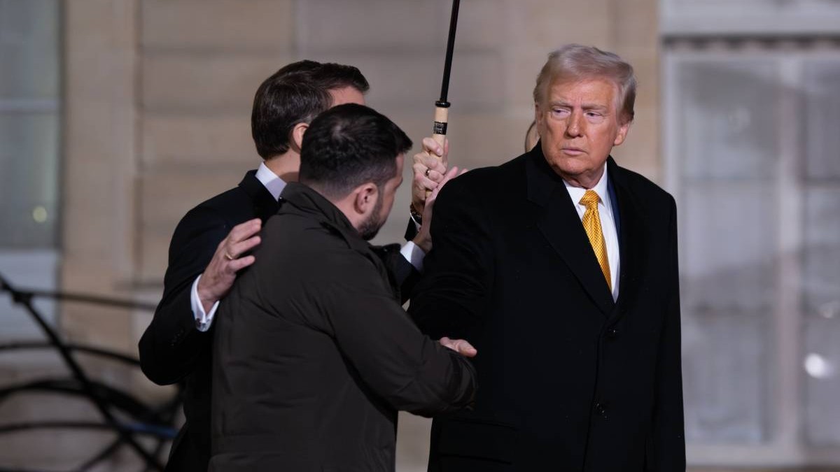 donald trump imbracat cu o camasa alba si palton negru in timp ce da mana cu volodimir zelenski. in spatele lor este emanuel macron