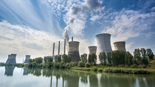 Termocentrala Turceni, cu un lac în față, turnurile uriașe pe fundal pe care iese fum și cerul albastru cu nori albi