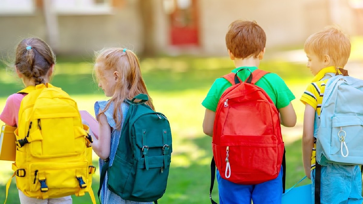 patru elevi cu ghiozdanele colorate în spate privasc spre terenul de joacă din fața lor