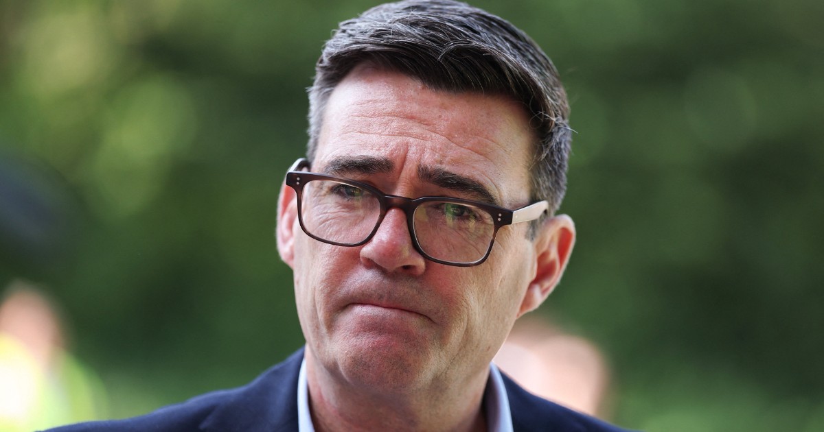 Imaginea îl surprinde pe Andy Burnham într-un prim-plan, purtând costum închis la culoare, cămașă deschisă și ochelari, cu o expresie serioasă și concentrată.