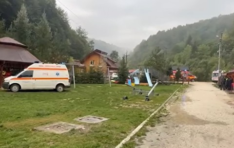 O ambulanță și alte vehicule de intervenție staționate în curtea unei cabane, înconjurată de munți
