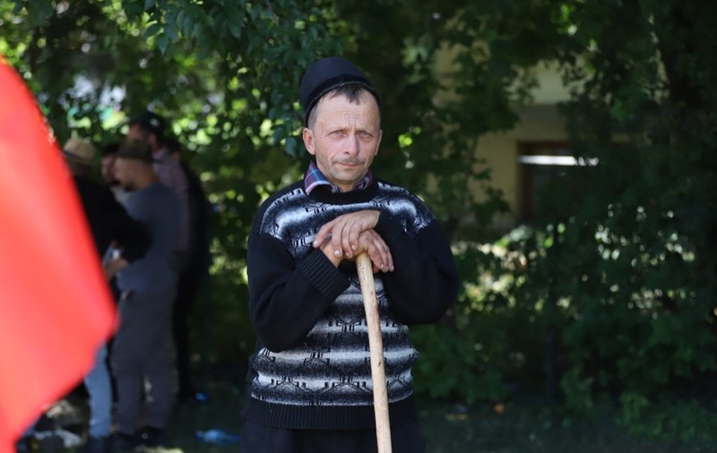 Ciobanul Liviu din Bihor, un bărbat cu un pulover albastru de lână, lângă un gard viu din București. Își ține capul sprijinit într-un toiag