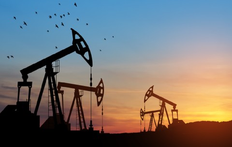 Imaginea prezintă mai multe sonde de petrol aflate în funcțiune pe fundalul unui apus de soare colorat, în timp ce un stol de păsări zboară pe cerul de deasupra lor.