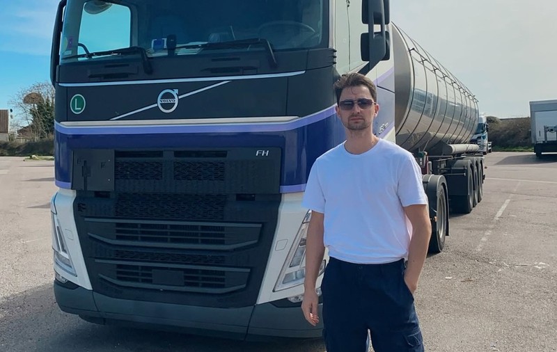 Un bărbat îmbrăcat în tricou alb, cu ochelari de soare, în fața unui camion cisternă Volvo într-o parcare.