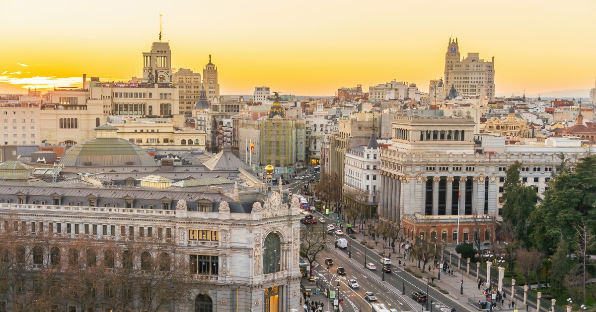 Imaginea surprinde o panoramă spectaculoasă a orașului Madrid la apus, cu cerul luminat în nuanțe aurii. În prim-plan se află clădiri istorice impunătoare, bulevarde largi și trafic intens, iar în fundal se conturează silueta modernă a capitalei spaniole.