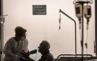 pacient stand pe pat intr-un salon de spital cu o perfuzie la capul patului. Langa el sta o femeie care-i pune masca de oxigen pe gura