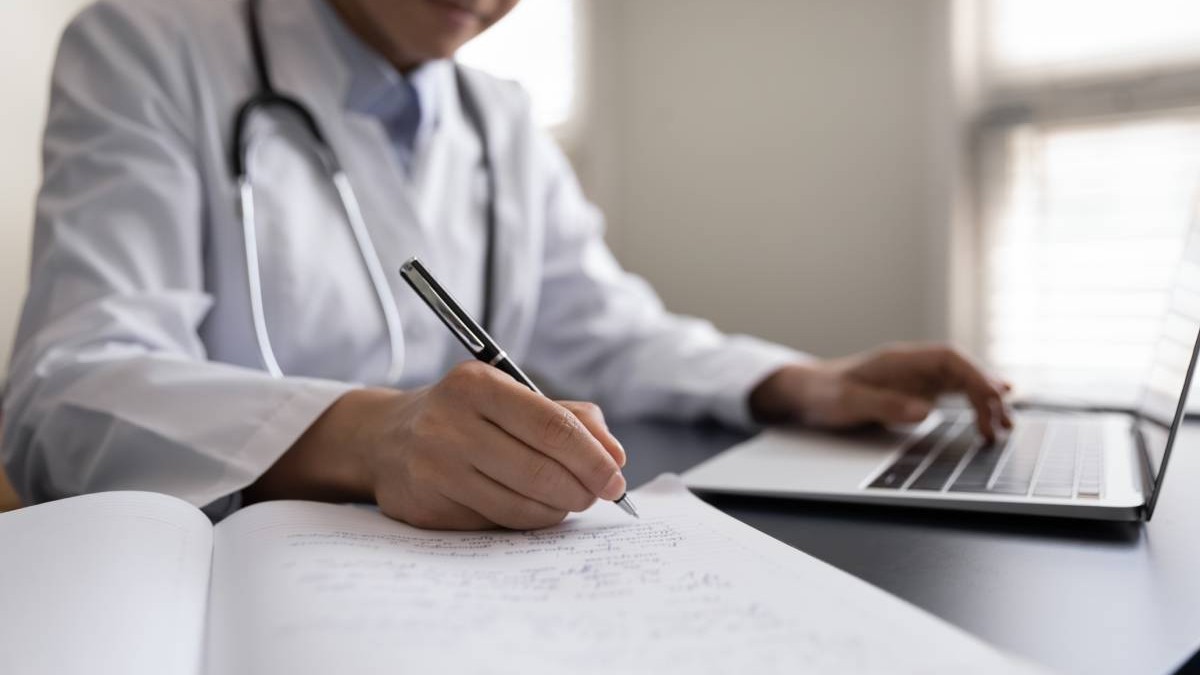 Un medic de familie, în timp ce stă la birou, în fața laptopului și notează datele pacientului asigurat la sănătate.