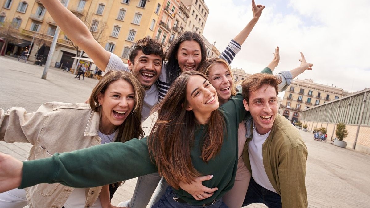Un grup de tineri prieteni entuziaști, care pozează fericiți pentru o fotografie în piața orașului. Tineri din Generația Z zâmbind cu brațele ridicate în aer pentru o fotografie de amintire din vacanță, bucurându-se de distracție într-un oraș european
