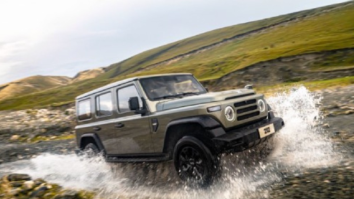 Imaginea prezintă o mașină de teren de culoare verde-măslinie, cu patru uși, în plină acțiune. Mașina, care seamănă cu un Jeep sau un Land Rover, traversează o apă curgătoare, iar apa țâșnește din lateral. Pe fundal se observă un peisaj deluros, cu vegetație scundă și un cer parțial înnorat.