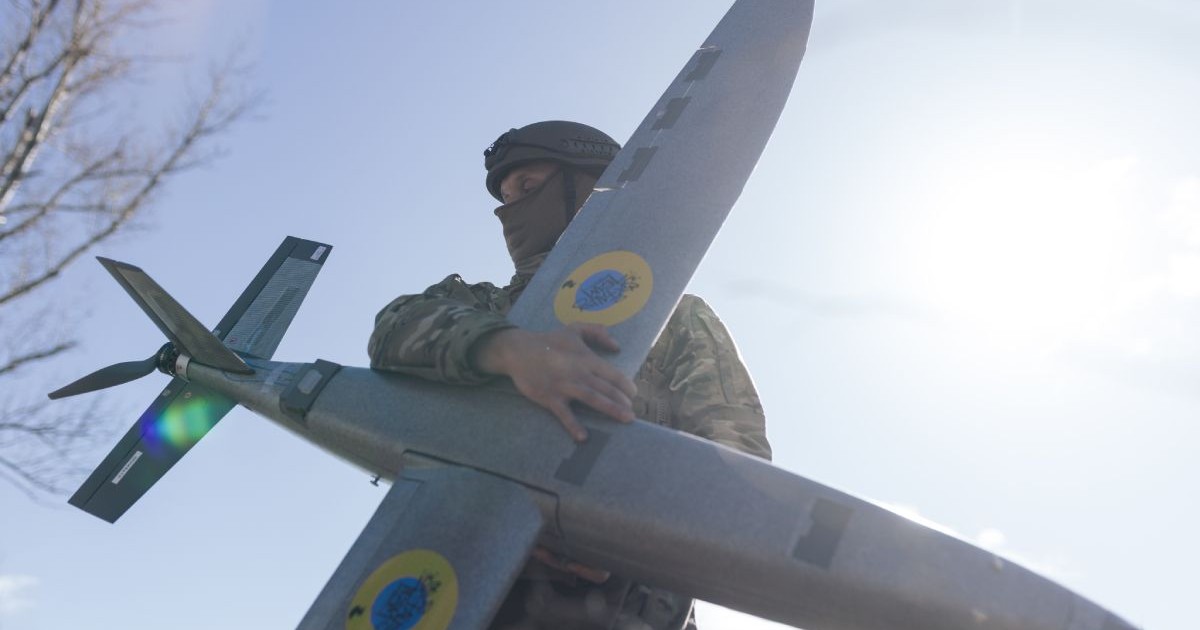 Un soldat ucrainean îmbrăcat în uniformă ține cu ambele mâini o dronă cu aripi, cu cerul albastru și Soarele pe fundal