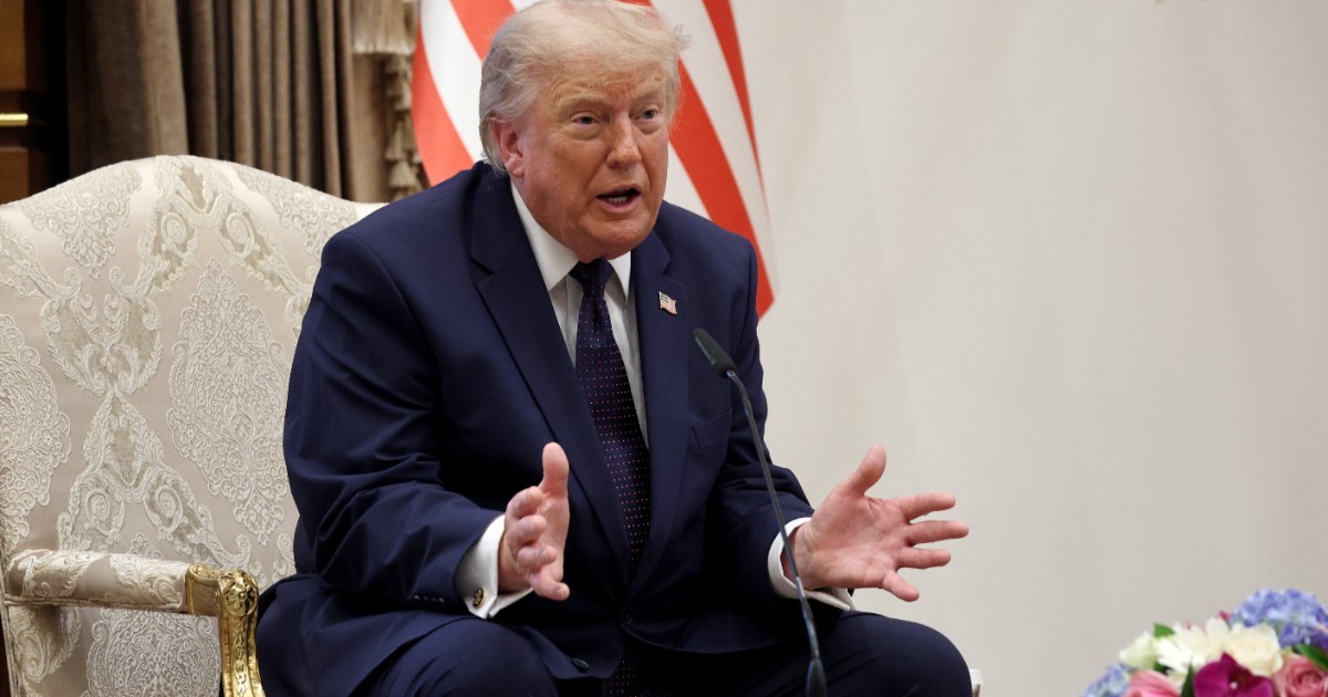 În imagine, Donald Trump este surprins vorbind și gesticulând cu ambele mâini în timp ce stă așezat pe un scaun, având steagul Statelor Unite în fundal