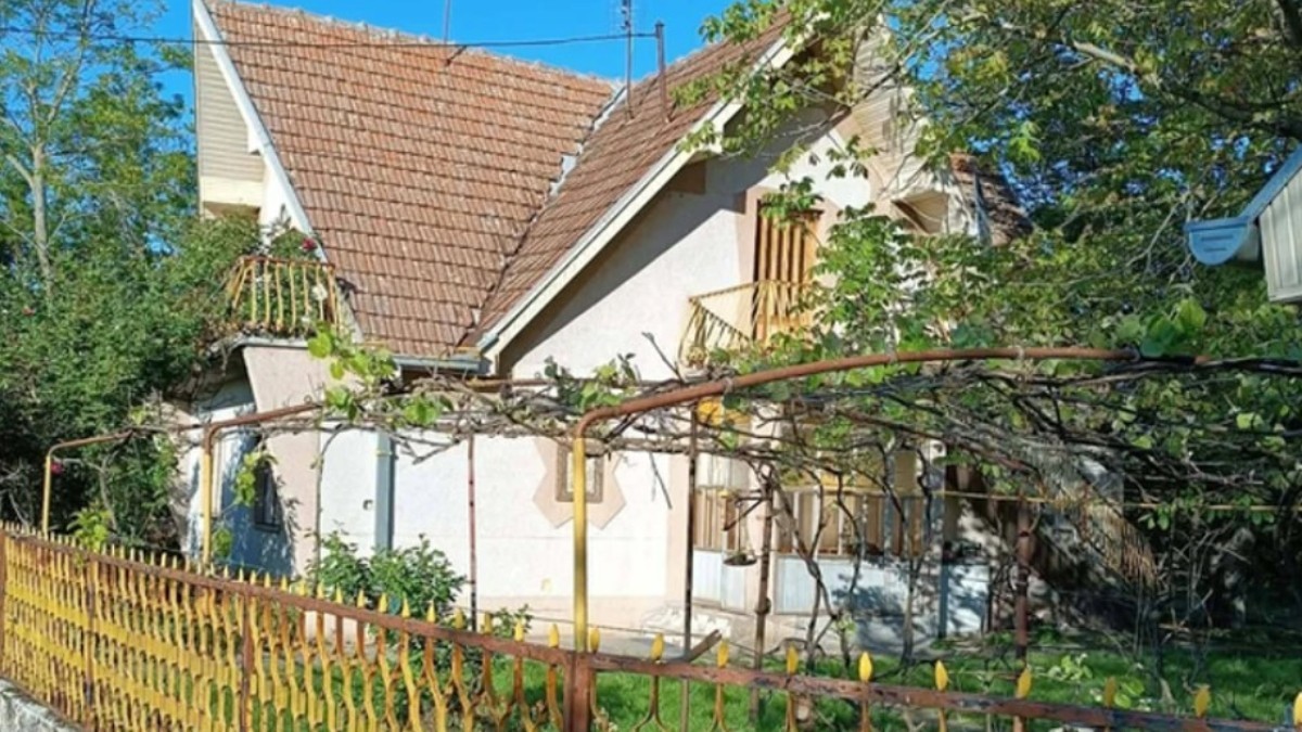 O casă cu etaj și mansardă, într-o curte plină de trandafiri. Are gardul galben și acoperișul din țiglă roșie