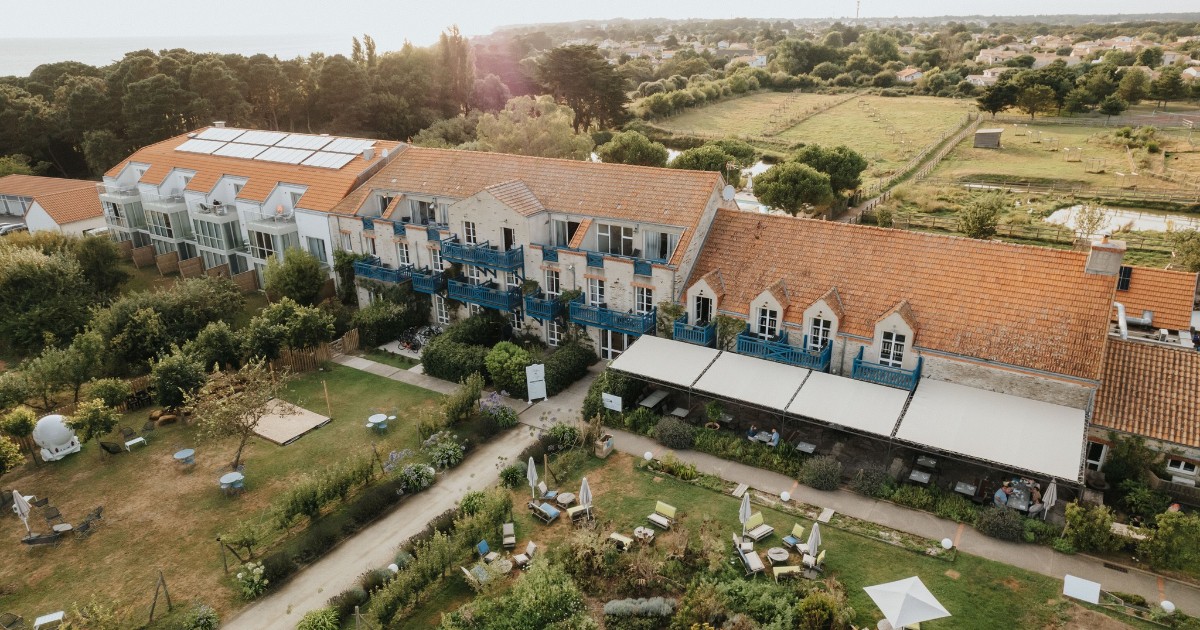 Imaginea surprinde un complex turistic amplasat într-un cadru natural, cu clădiri cu acoperișuri roșii și panouri solare instalate pe acoperiș. În jur se observă grădini amenajate, spații verzi și zone de relaxare, sugerând un loc dedicat turismului sustenabil și liniștii.