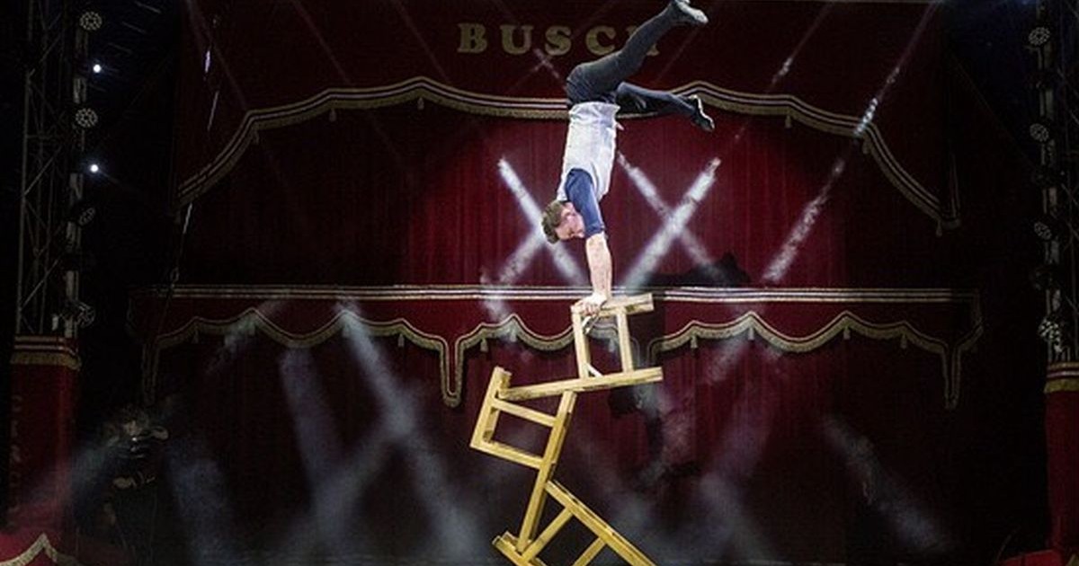 Un acrobat în timpul unui spectacol de circ se ține în mâini pe niște scaune așezate pe o masă și este cu picioarele în sus