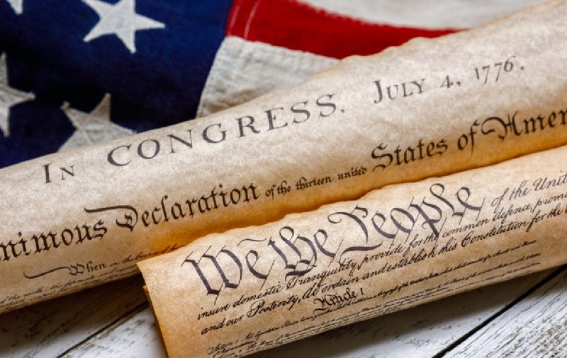 Exemplare înfășurate ale Constituției SUA și ale Declarației de Independență, alături de un steag american de epocă, pe lemn vopsit în alb și cu aspect patinat