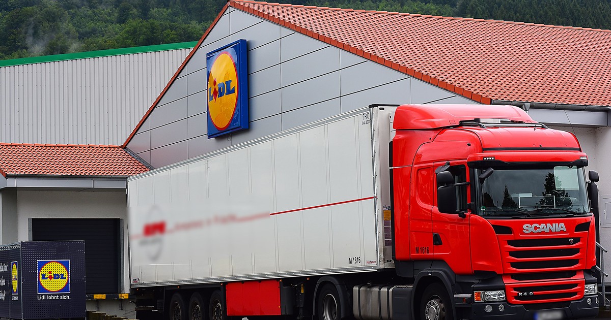 Imagine ilustrativă cu un camion Scania roșu cu remorcă albă parcat la rampa de încărcare a unui supermarket Lidl. Fundalul include un acoperiș cu țiglă roșie, copaci verzi și un cer înnorat