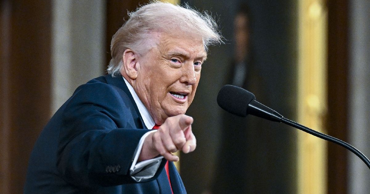 Președintele Donald Trump, îmbrăcat într-un costum bleumarin și cravată roșie, arătând cu degetul spre aparatul de fotografiat, în fața unui microfon.