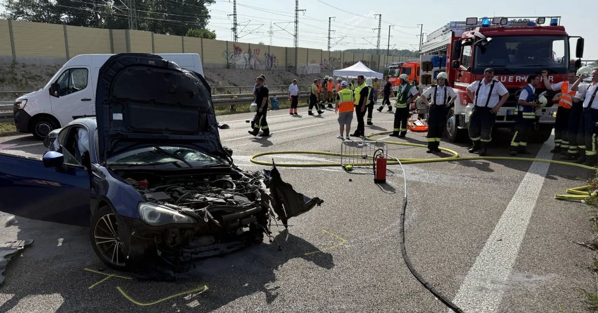 În prim-plan este o mașină neagră Subaru, cu capota ridicată și lovită, după ce i-a explodat un cauciuc, în spatele ei este o dubiță albă, iar în spate este o autospecială de pompieri și mai mulți oameni la fața locului, accident Germania, 14 iunie 2025