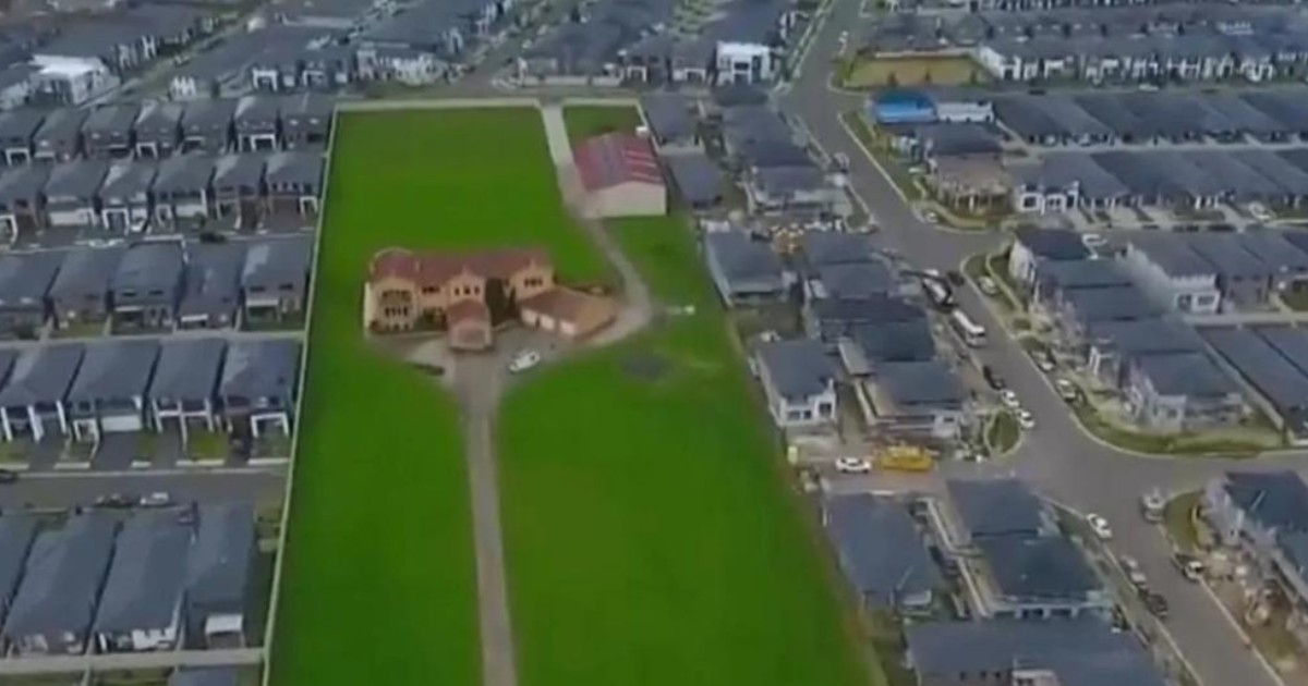 În imagine se vede o casă amplasată pe un teren foarte mare, cu iarbă verde, rămasă izolată în mijlocul unui cartier modern și dens construit. În jurul proprietății, casele sunt lipite unele de altele, în timp ce locuința veche păstrează o zonă vastă liberă, contrastând puternic cu restul zonei urbane.