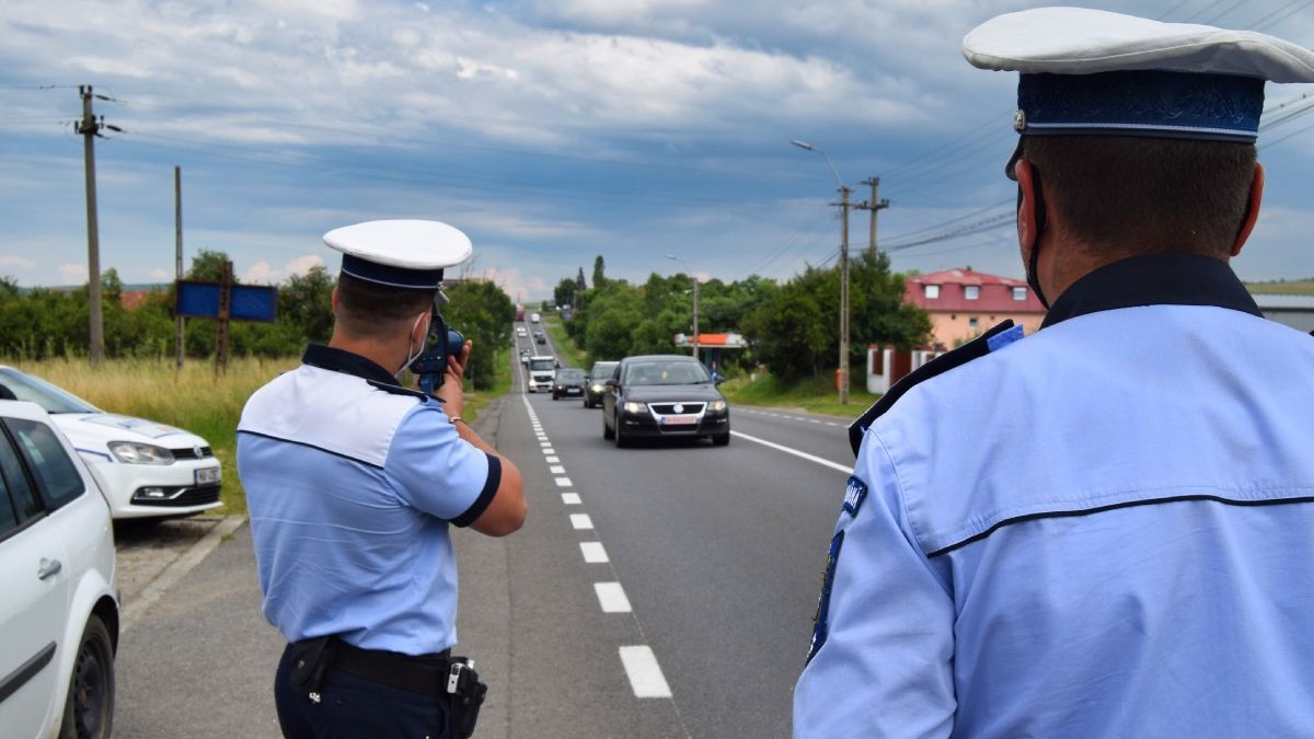 Doi polițiști îmbrăcați în uniforme, cu cămași albastre cu mâneci scurte, unul în prim plan, altul în plan îndepărtat care ține în mână un radar de tip pistol și filmează traficul pe o șosea pe care circulă mașini, cu cerul albastru pe fundal