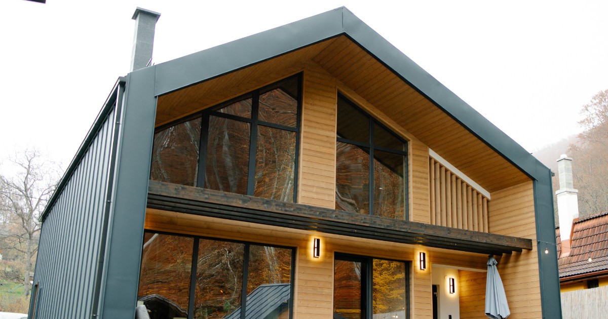Imaginea prezintă o casă modernă cu două niveluri, construită din lemn și metal, cu ferestre mari din sticlă care oferă multă lumină naturală. Locuința are o terasă în față și este amplasată într-un cadru liniștit, înconjurat de natură și copaci.