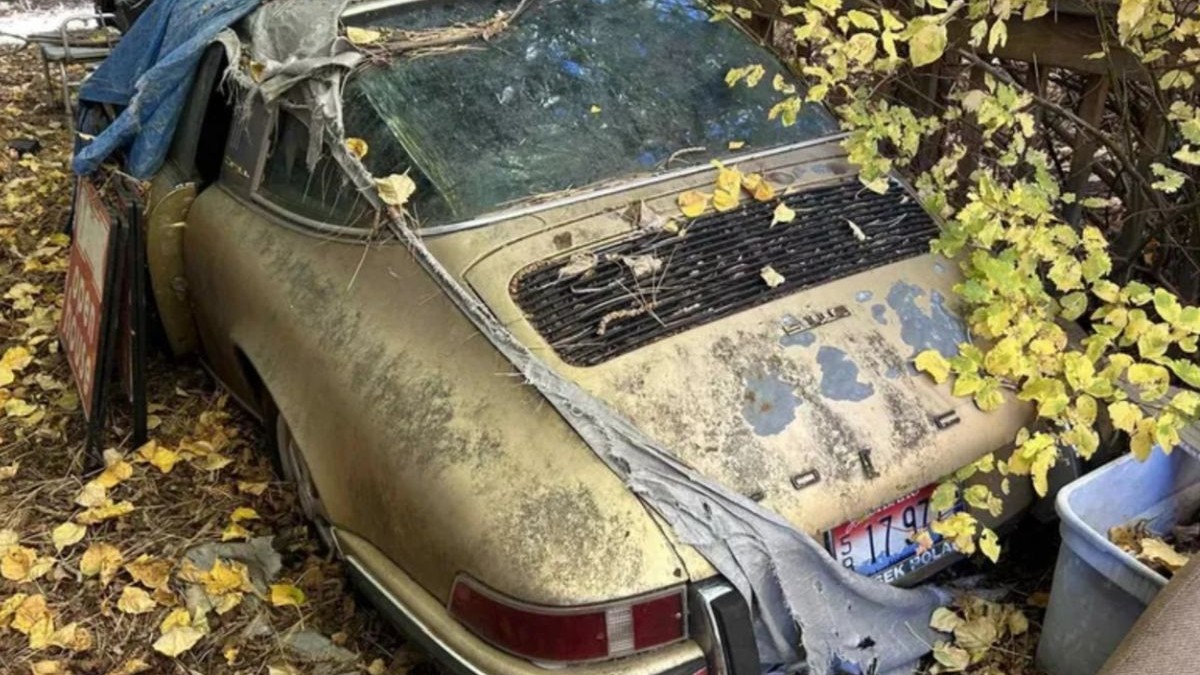 O mașină Porsche 911, fotografiată din spate. Se vede cum vopseaua este cojită și sărită în unele locuri. O copertină veche acoperă partea din față a autoturismului. Frunzele galbene de toamnă sunt căzute peste mașină