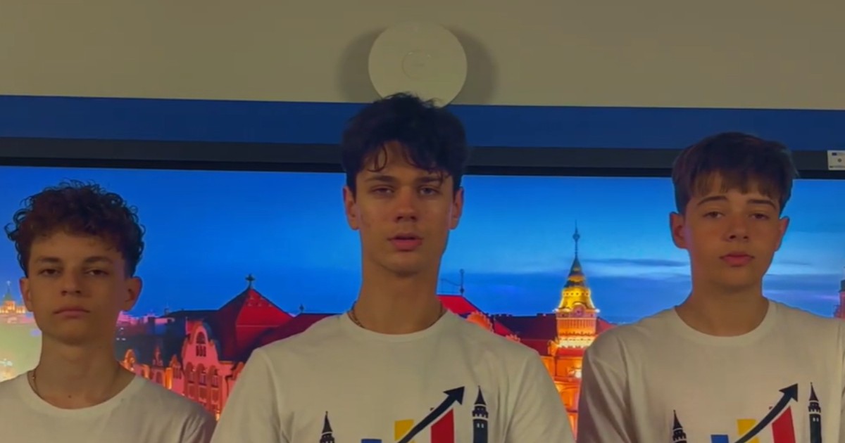 Trei adolescenți poartă tricouri albe cu un logo care include un grafic ascendent și culorile drapelului României. Ei stau în fața unui ecran pe care este afișată o imagine urbană luminată, părând să participe la o prezentare sau la un proiect educațional.