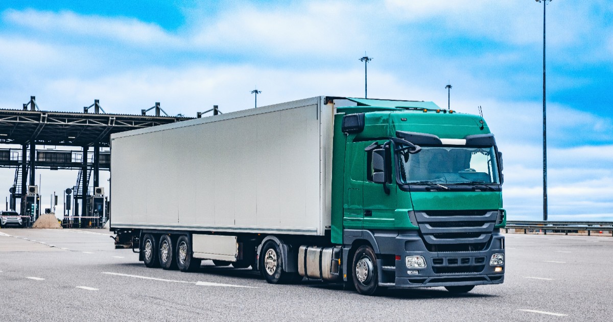 Un autocamion de mare tonaj, cu o cabină de culoare verde și o remorcă albă, circulă pe un carosabil asfaltat sub un cer parțial înnorat, după ce a trecut de o zonă de control.