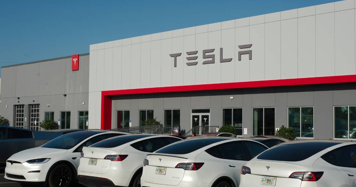 Exteriorul unei cladiri Tesla, un showroom sau un service auto. În fața clădirii sunt parcate mai multe vehicule Tesla albe, în special modele Model Y și Model X