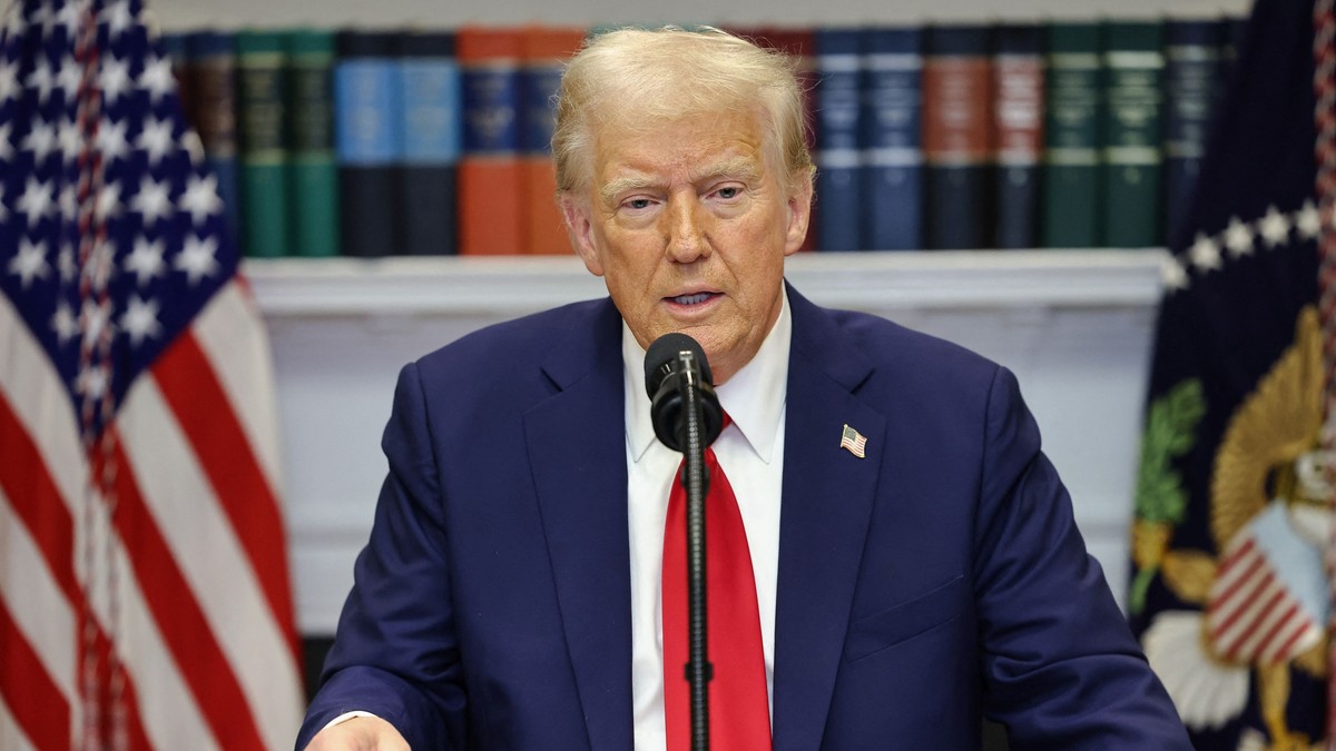 Donald Trump, într-o conferință de presă la Casa Albă, îmbrăcat într-un costum bleumarin cu o cravată roșie, așezat în fața steagului Americii