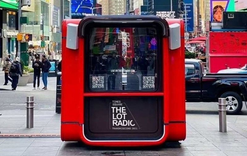 Un chioșc roșu de studio radio pop-up al postului „The Lot Radio”, instalat în spațiul public din Times Square, New York, având difuzoare pe margini și panouri cu coduri QR