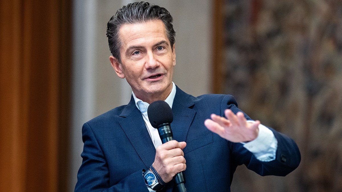 Roland Weissmann cu o cămașă deschisă și la culoare și sacuo ănchis la culaore vorbește la microfon.