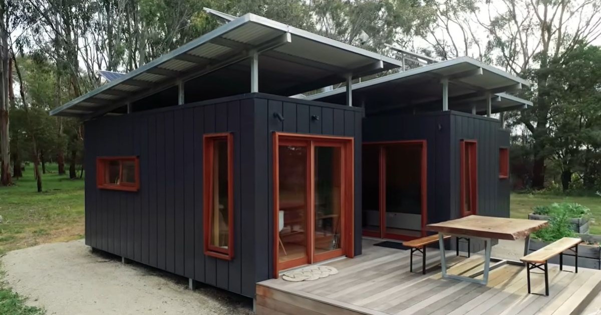 Casa construită din containere, de culoare negru, cu o terasă de lemn în față, iar pe fundal o pădure verde