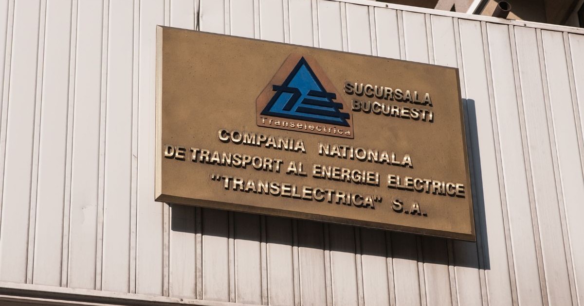 plăcuță metalică afișată pe fațada unei clădiri cu logo-ul companiei de stat Transelectrica și textul „Compania Națională de Transport al Energiei „Tranelectrica” S.A., scris cu litere metalice aurii pe un fundal maro