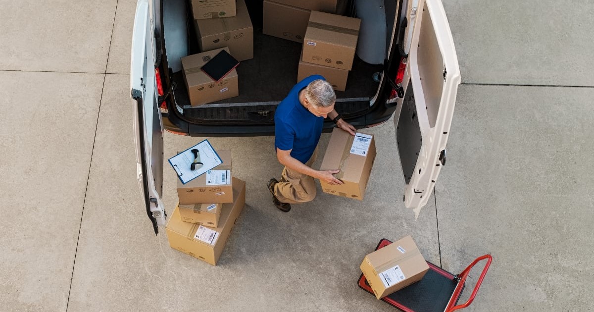 Vedere de sus a unui curier în tricou albastru care descarcă pachete din carton din spatele unei furgonete albe cu ușile deschise, având lângă el alte cutii stivuite și un cărucior manual de transport