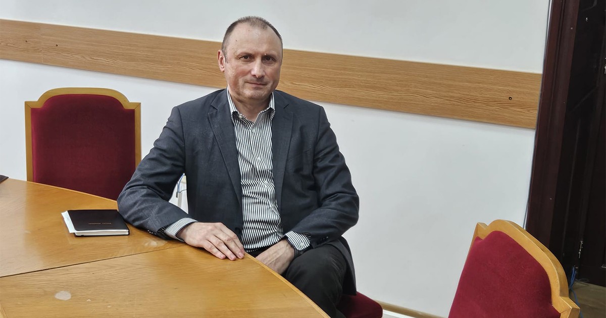 Un barbat in costum gri sta la o masa si acordă un interviu