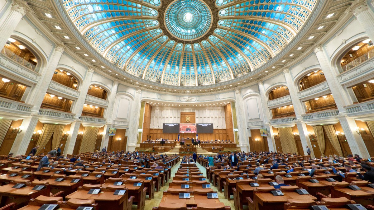 Imaginea prezintă o vedere de ansamblu din interiorul plenului Camerei Deputaților din Palatul Parlamentului, cu un vitraliu albastru pe cupolă și băncile parlamentarilor pregătite pentru ședință.