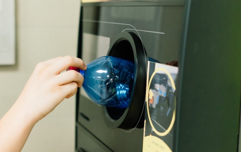 O mână care introduce o sticlă din plastic într-un aparat automat pentru colectarea și reciclarea ambalajelor.