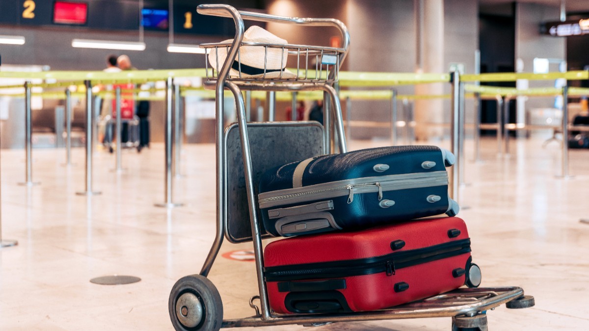 Un cărucior metalic de bagaje aflat într-un terminal de aeroport, încărcat cu două valize și o pălărie
