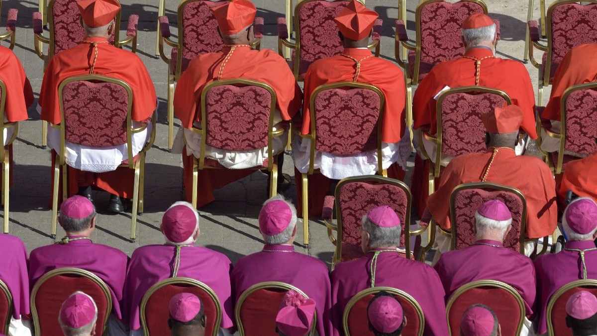 Mai mulți cardinali ai Bisericii Catolice, îmbrăcați în roșu sau roz, stau așezați pe scaune roșii