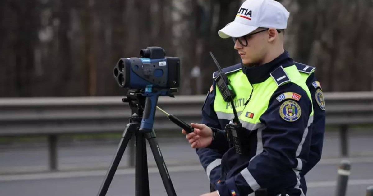 Polițist rutier echipat cu uniforma specifică și o șapcă albă inscripționată cu „POLIȚIA”. Acesta operează un radar mobil de tip pistol montat pe un trepied. Pe fundal se observă un drum național, delimitat de un parapet metalic.