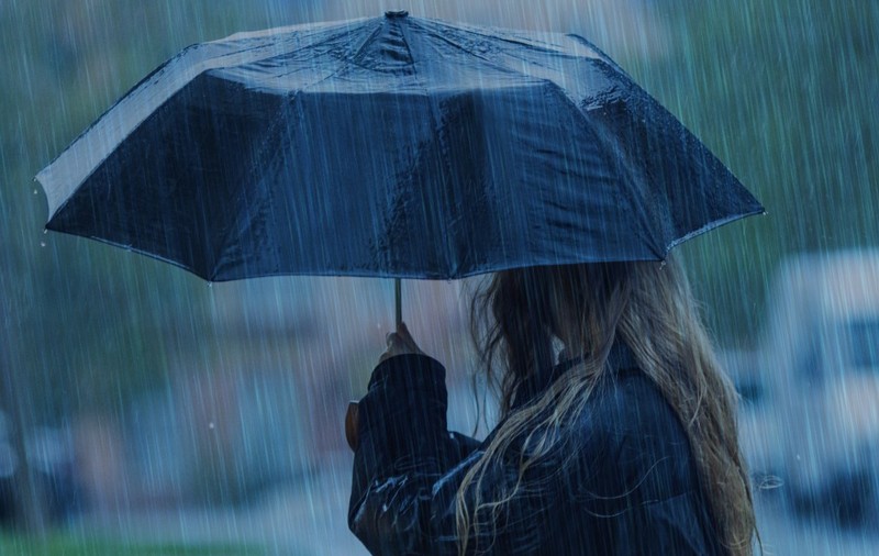 O persoană cu părul lung și blond, privită din lateral, care ține o umbrelă neagră deschisă pe o ploaie torențială.