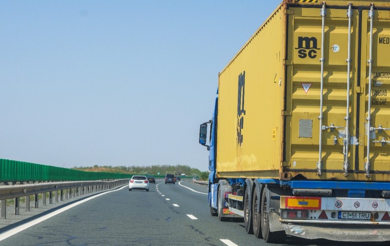 Imaginea surprinde un camion albastru ce transportă un container galben, rulând pe o autostradă alături de alte autoturisme, sub un cer senin.