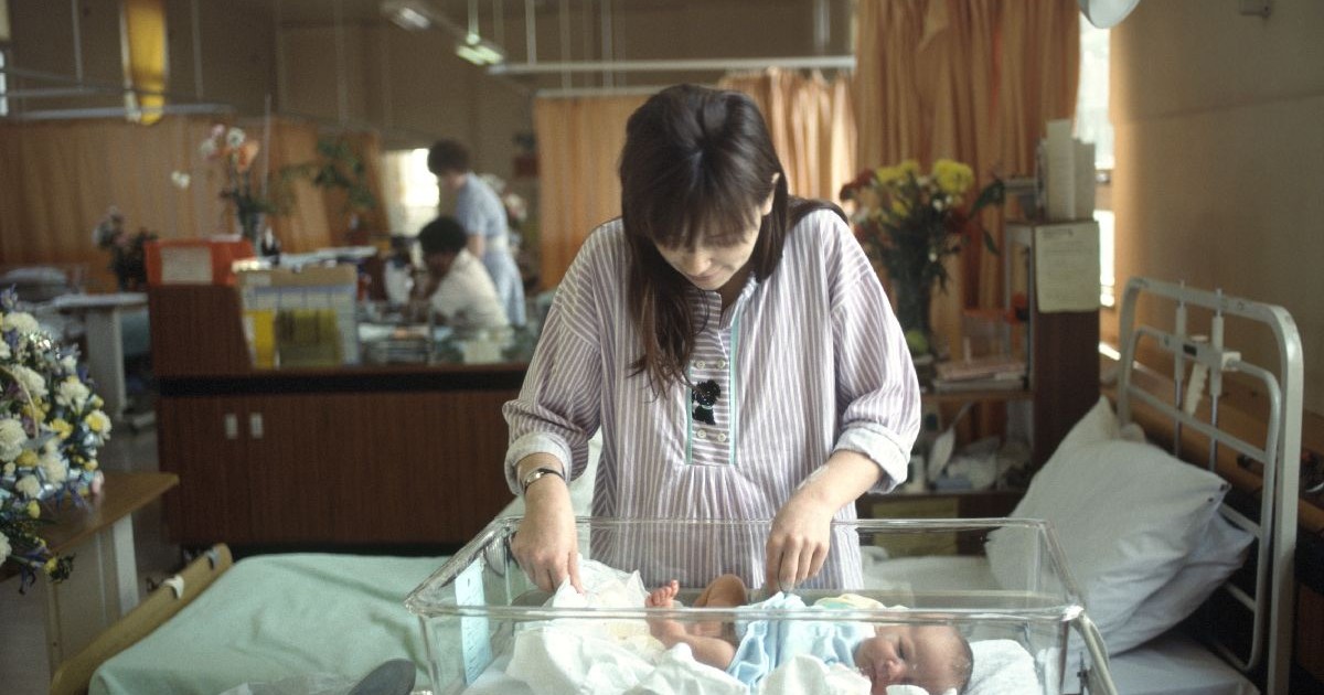 O mama stă în picioare lângă copilul ei nou născut, într-un salon de spital