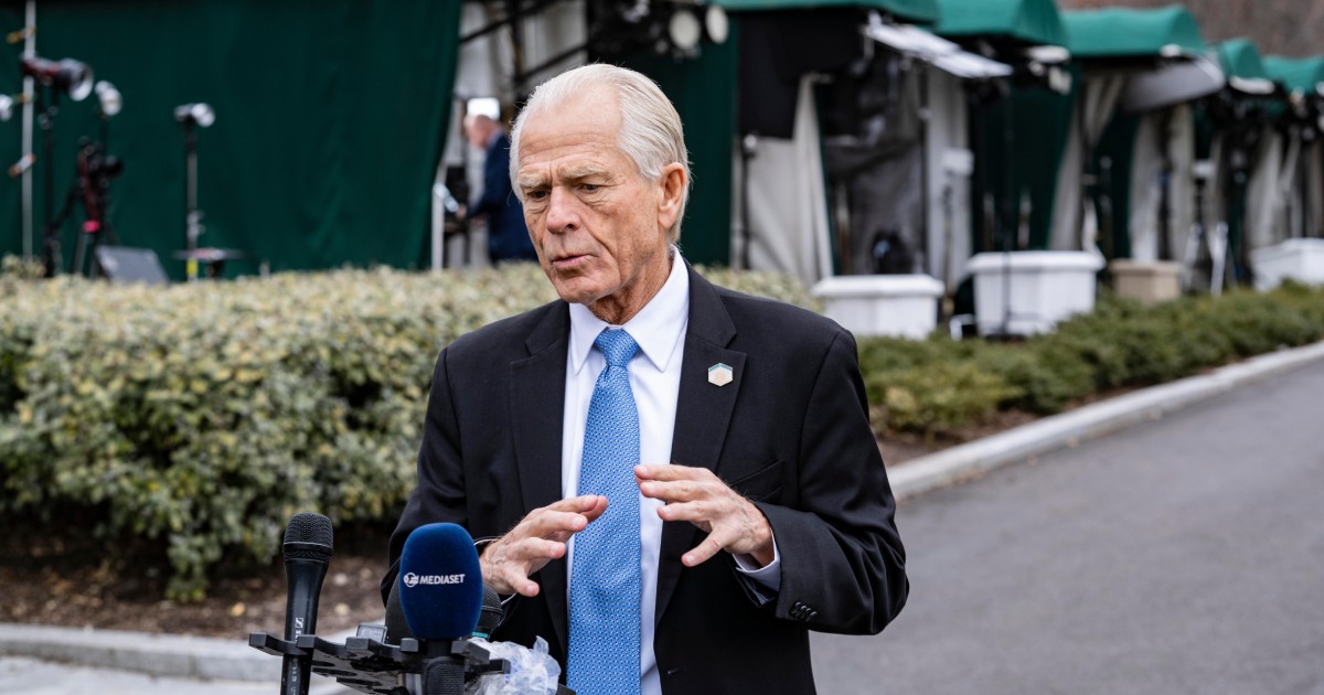 Peter Navarro, îmbrăcat într-un costum negru, cămașă albă și cravată albastră. Are părul alb și gesticulează cu ambele mâini în timp ce vorbește la microfon, afară, la o conferință de presă
