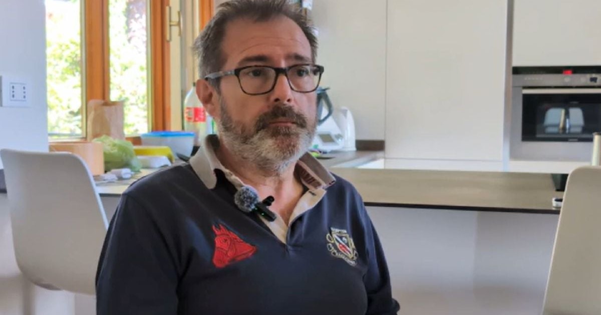 Imaginea surprinde un bărbat cu ochelari și barbă grizonată, purtând un tricou albastru închis cu o lavalieră atașată, în timp ce pare să ofere un interviu.