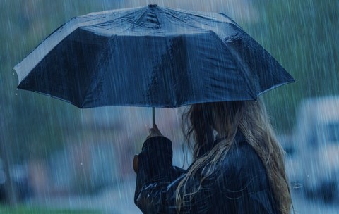 O persoană cu părul lung și blond, privită din lateral, care ține o umbrelă neagră deschisă pe o ploaie torențială.
