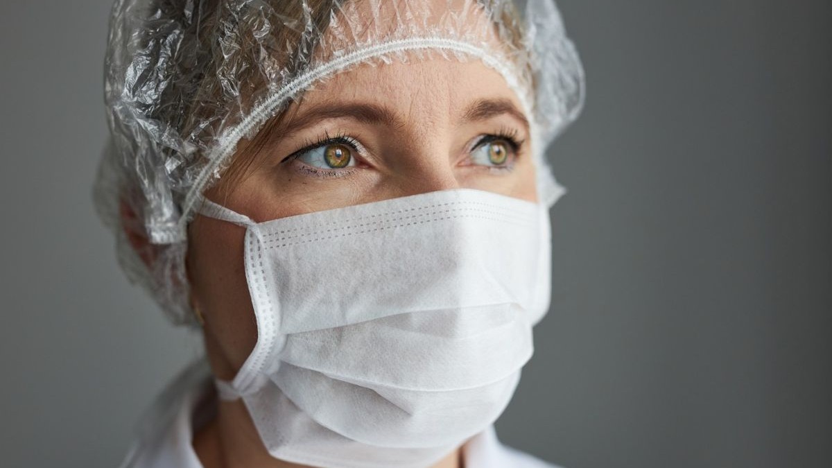 O doctoriță cu fața acoperită cu mască de protecție și capul acoperit, într-un cabinet medical, cu un perete gri în spate