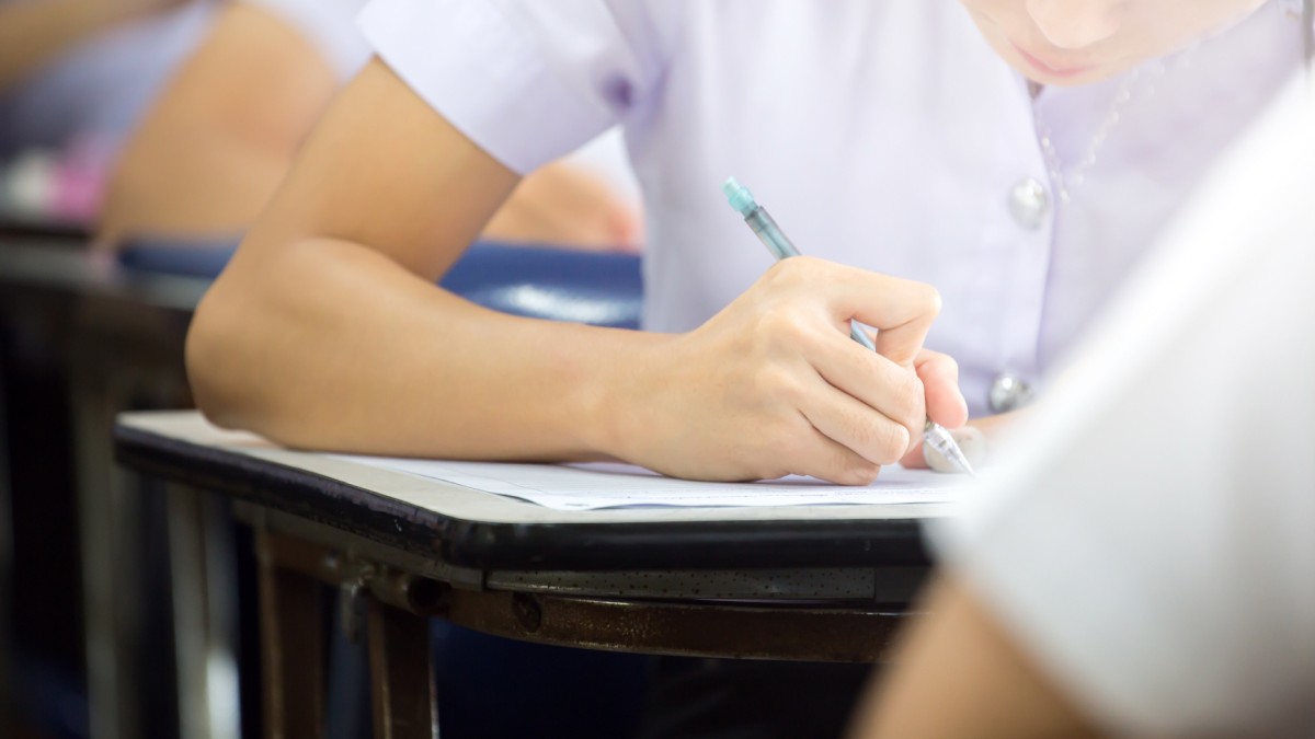 Mai muți elevi, surpinși de la gât în jos, stau în bănci unul în spatele celuilalt, și scriu pe foi de hârtie, în timpul unui examen.